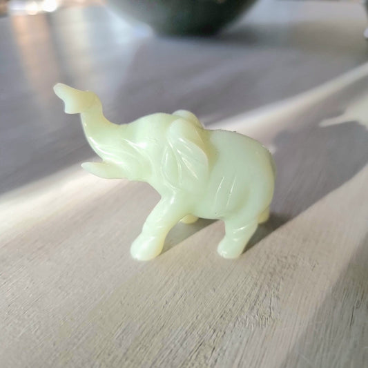 Small carved pale green stone elephant figurine with raised trunk, standing pose, on a light wooden surface in natural light