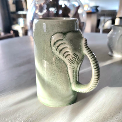 Thai celadon crackle glaze ceramic elephant mug with elephant head and trunk forming the handle, soft green with weblike crackle pattern, on a pale surface