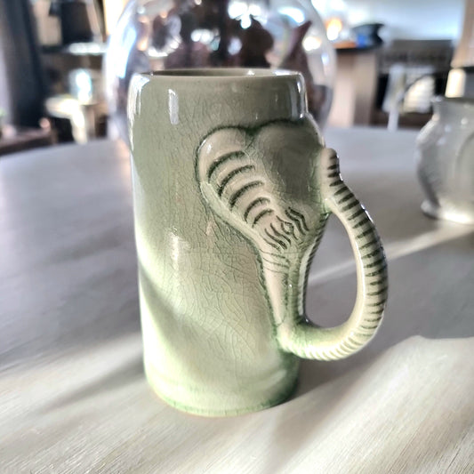 Thai celadon crackle glaze ceramic elephant mug with elephant head and trunk forming the handle, soft green with weblike crackle pattern, on a pale surface