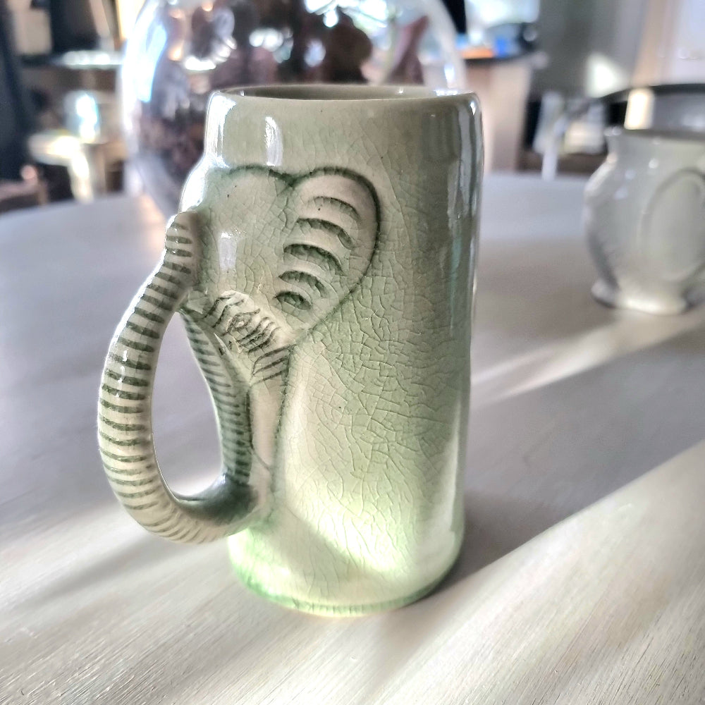 Thai celadon crackle glaze ceramic elephant mug with elephant head and trunk forming the handle, soft green with weblike crackle pattern, on a pale surface