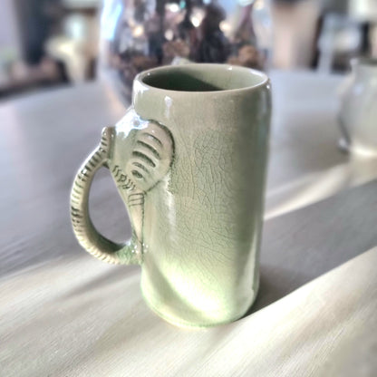 Thai celadon crackle glaze ceramic elephant mug with elephant head and trunk forming the handle, soft green with weblike crackle pattern, on a pale surface