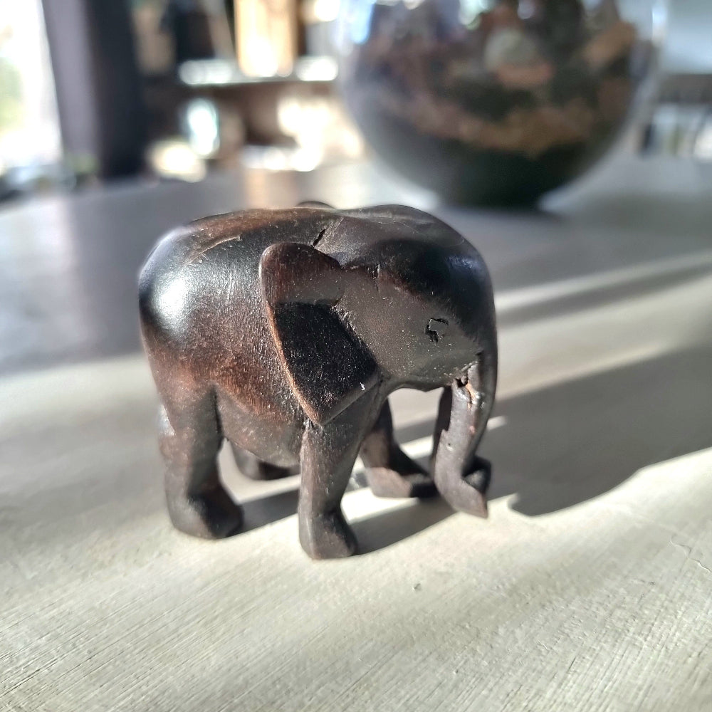 Small hand-carved dark ebony-toned hardwood elephant figurine with tusks and lowered trunk, standing pose, on a pale wooden surface in natural light