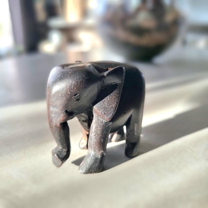 Small hand-carved dark ebony-toned hardwood elephant figurine with tusks and lowered trunk, standing pose, on a pale wooden surface in natural light
