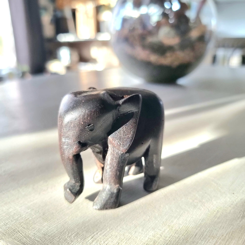 Small hand-carved dark ebony-toned hardwood elephant figurine with tusks and lowered trunk, standing pose, on a pale wooden surface in natural light