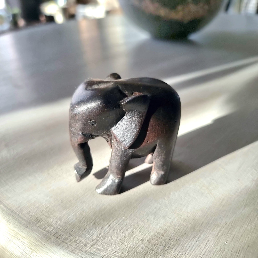 Small hand-carved dark ebony-toned hardwood elephant figurine with tusks and lowered trunk, standing pose, on a pale wooden surface in natural light