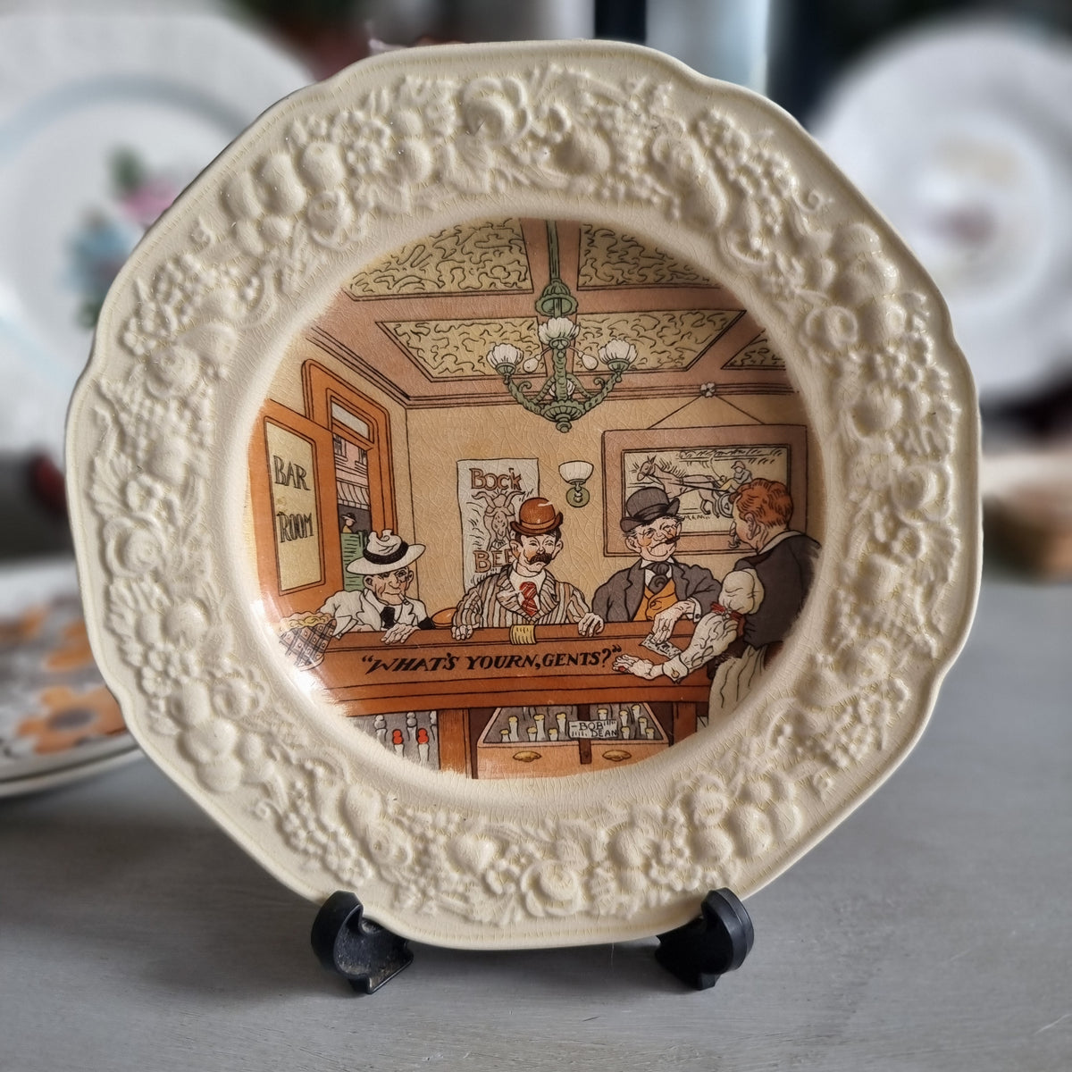 Vintage Crown Ducal Florentine plate showing a bar scene with men in hats and the words What's yourn, gents, in a moulded cream border