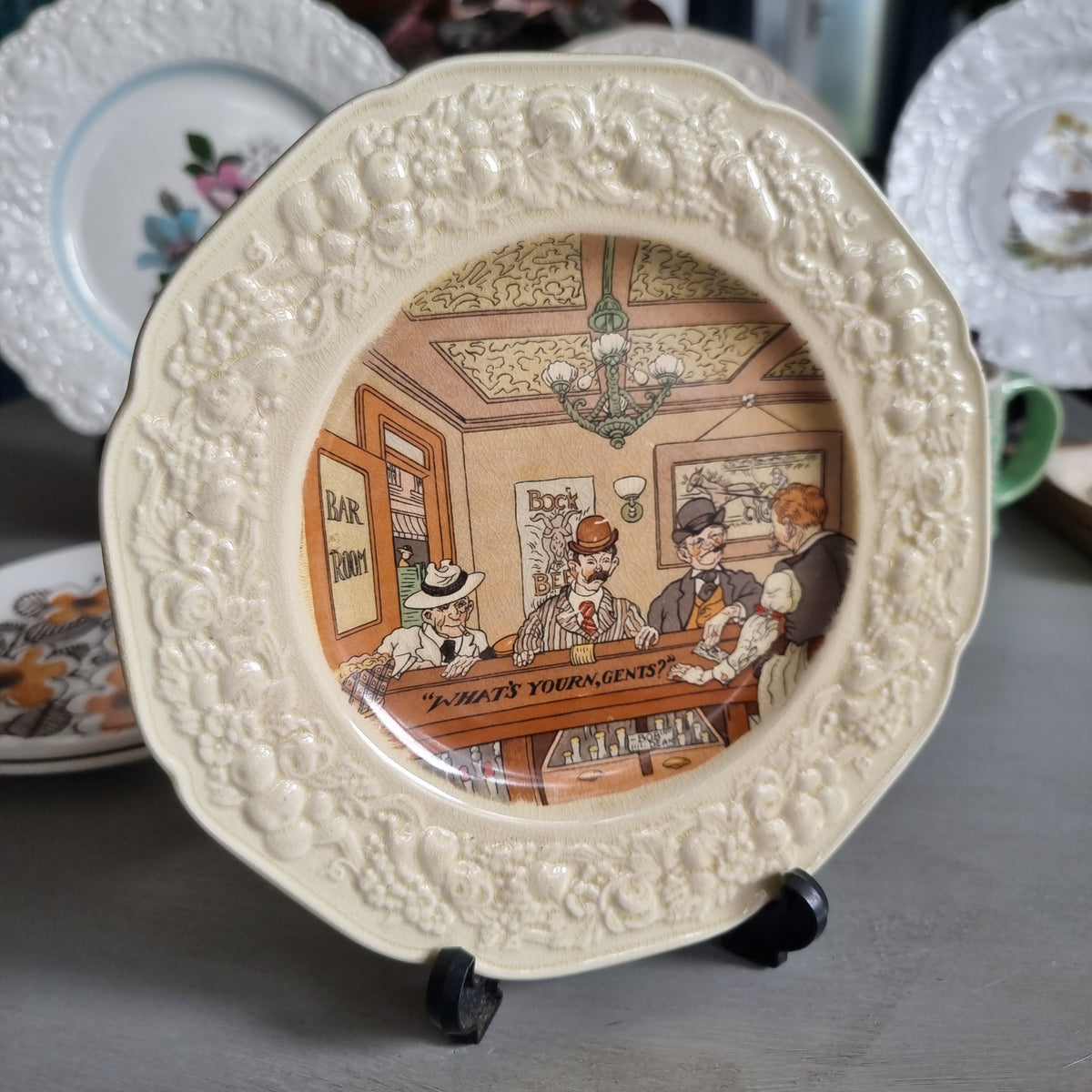 Vintage Crown Ducal Florentine plate showing a bar scene with men in hats and the words What's yourn, gents, in a moulded cream border