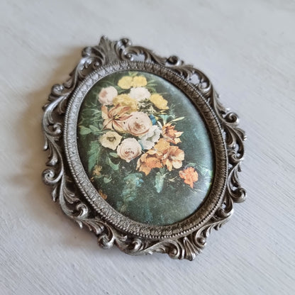 Small oval ornate pewter-tone frame holding a silk print of peach and cream roses on a dark green background