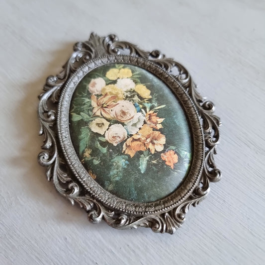 Small oval ornate pewter-tone frame holding a silk print of peach and cream roses on a dark green background
