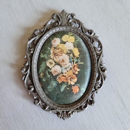 Small oval ornate pewter-tone frame holding a silk print of peach and cream roses on a dark green background