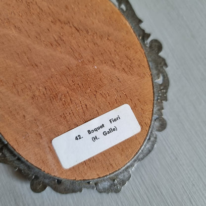 Small oval ornate pewter-tone frame holding a silk print of peach and cream roses on a dark green background