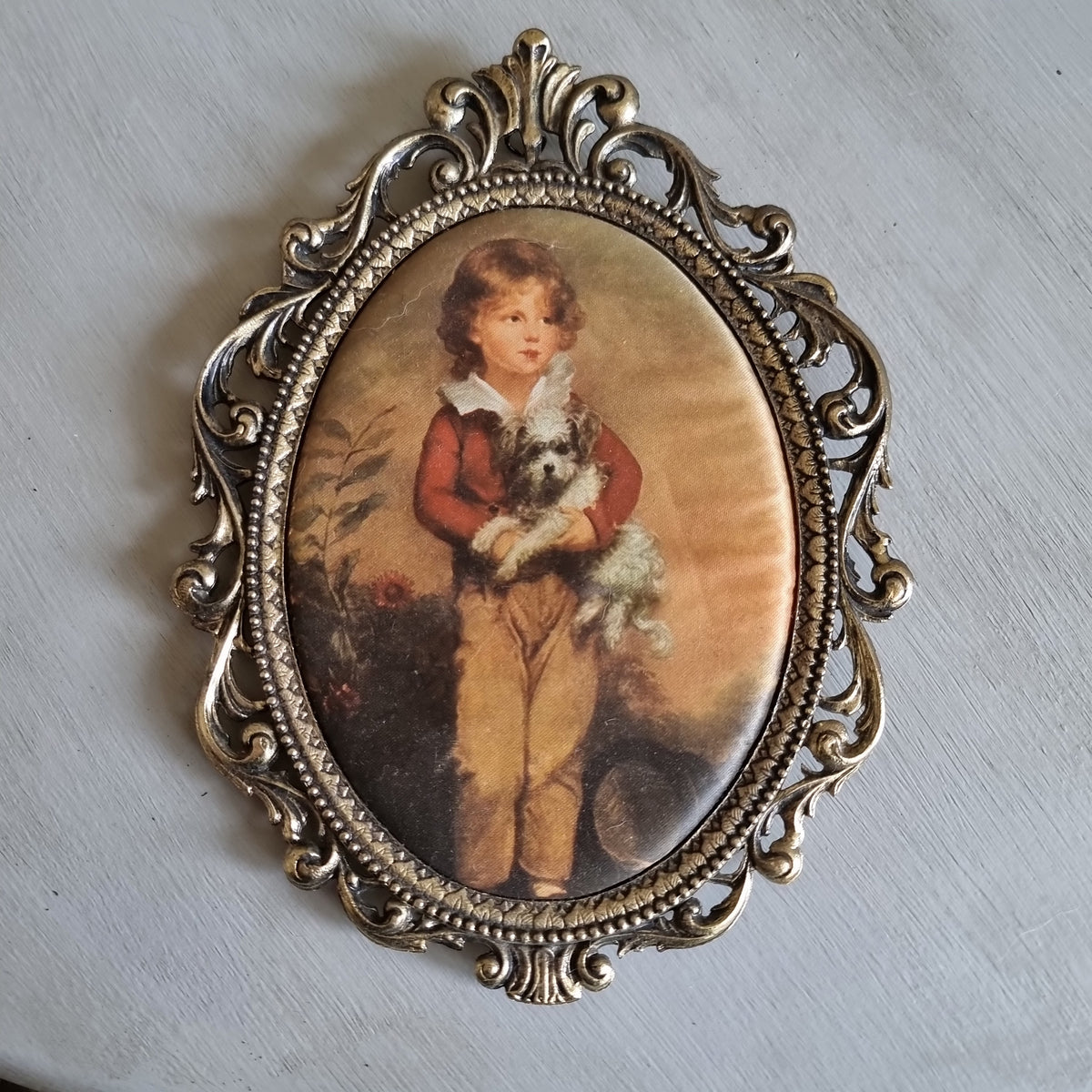 Ornate gold-tone brass oval frame with a silk print of a boy in a red jacket holding a small white dog