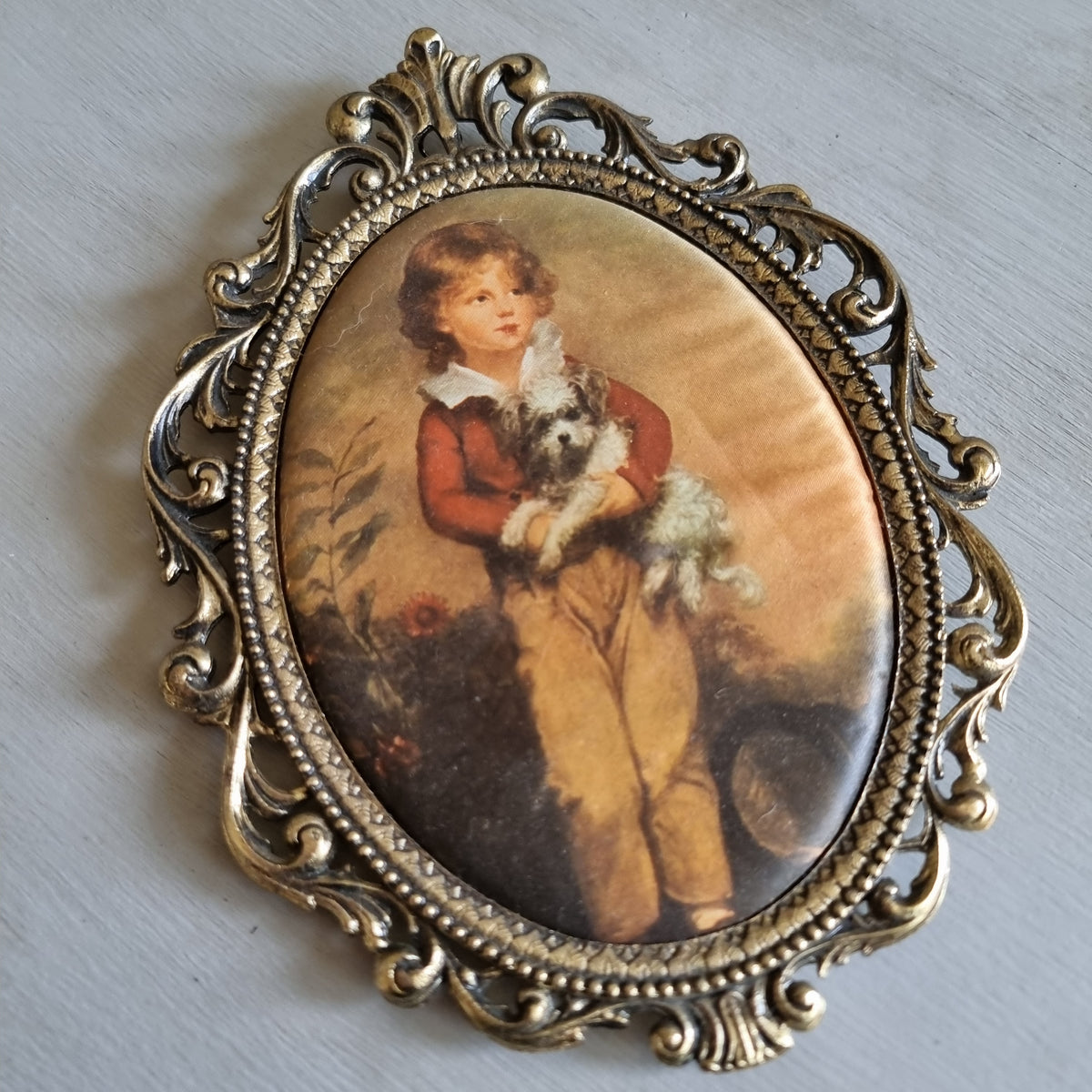Ornate gold-tone brass oval frame with a silk print of a boy in a red jacket holding a small white dog