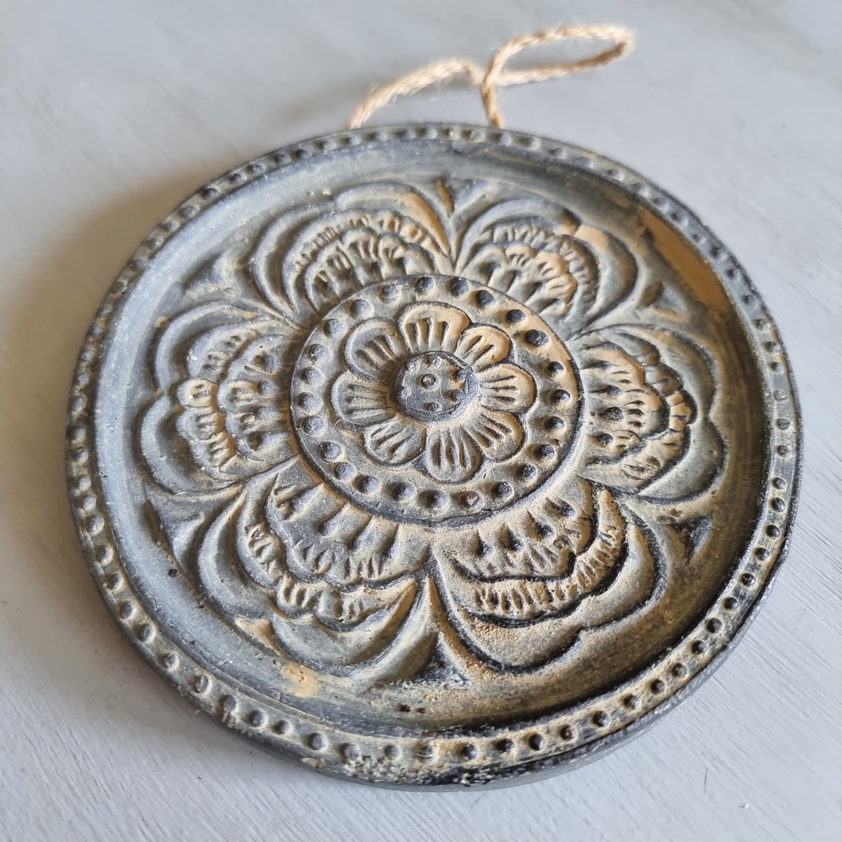 Round charcoal ceramic wall plaque with an embossed layered flower design highlighted in gold, hanging from a jute string