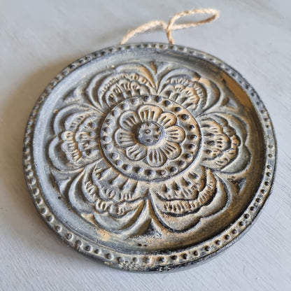 Round charcoal ceramic wall plaque with an embossed layered flower design highlighted in gold, hanging from a jute string