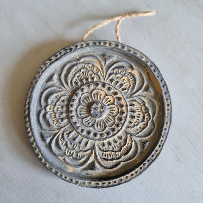 Round charcoal ceramic wall plaque with an embossed layered flower design highlighted in gold, hanging from a jute string