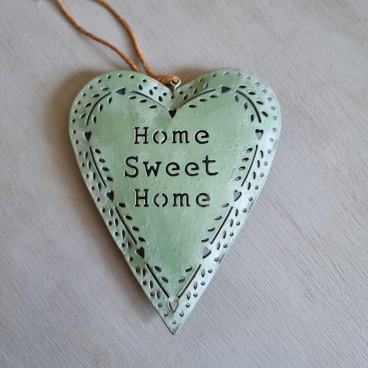 Sage green metal heart with Home Sweet Home cut out in the centre and a punched heart and leaf border, hanging from twine