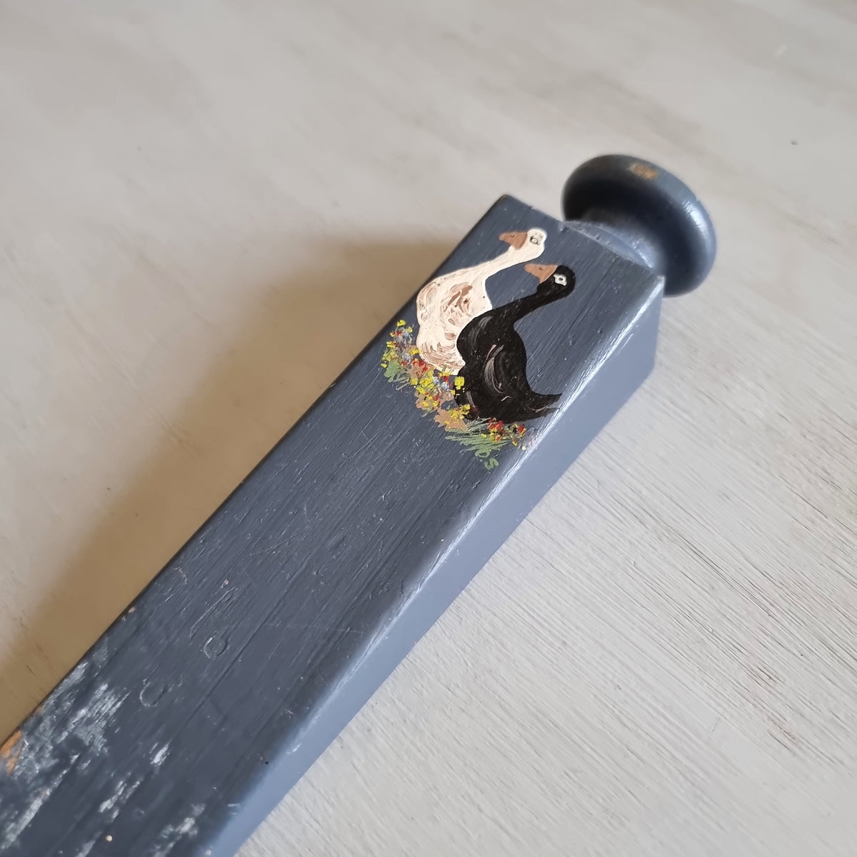 Grey painted wooden wedge door stop with a round knob, hand painted with a white goose and a black goose among flowers